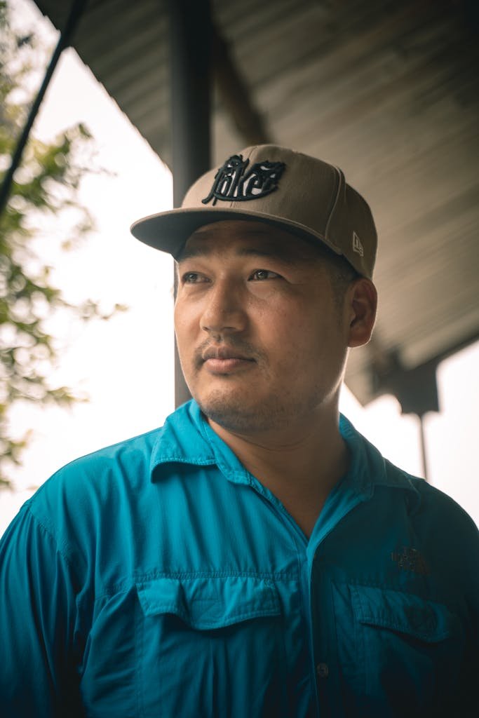 Portrait of a man wearing a blue shirt and cap, looking thoughtfully outdoors under a canopy.